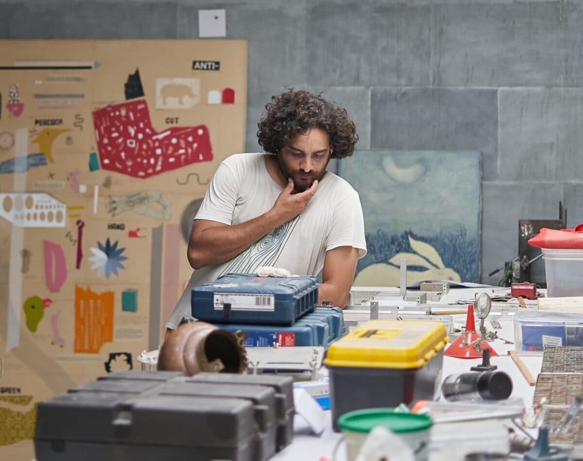 Eine Person mit lockigen Haaren steht nachdenklich an einem Tisch voller Werkzeuge, Kisten und Materialien. Im Hintergrund hängen bunte Poster und Bilder an der Wand.