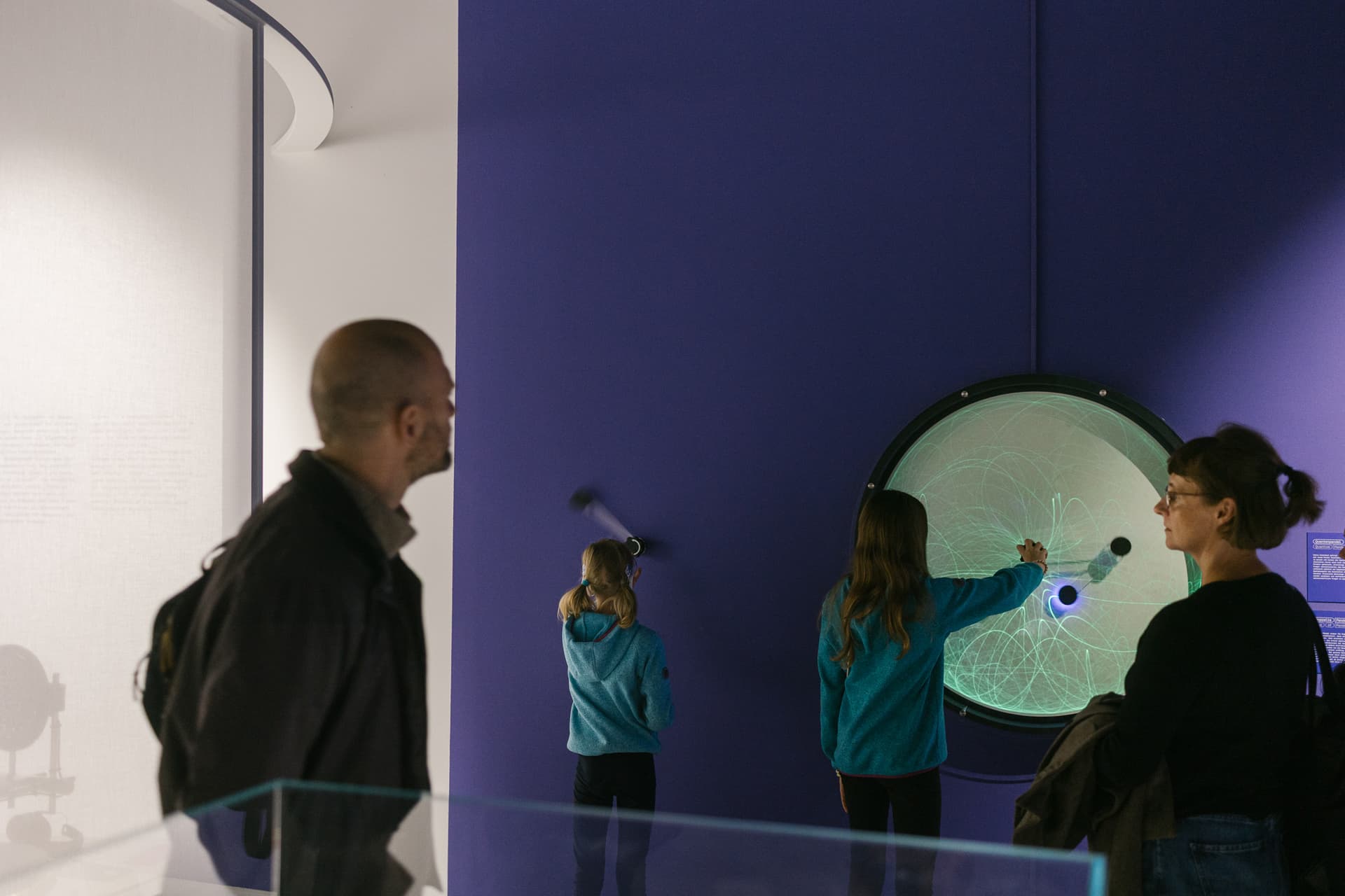 Menschen in einem Museum betrachten eine große interaktive Ausstellung an einer violetten Wand; zwei Kinder beschäftigen sich mit dem Exponat.
