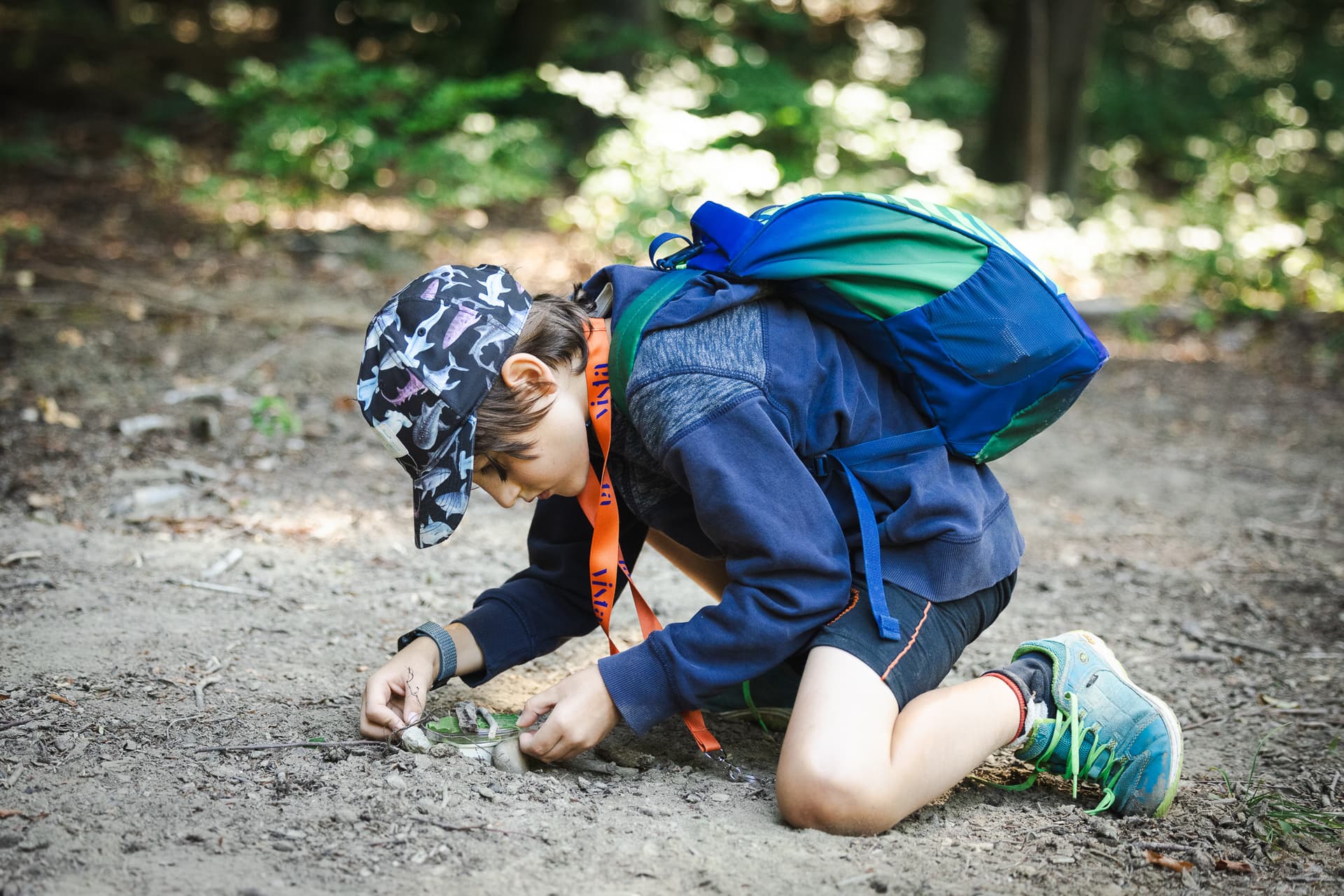 Kind kniet auf einem Waldweg und untersucht Blätter und Stöcke, trägt eine gemusterte Mütze, einen blaugrünen Rucksack und Turnschuhe.