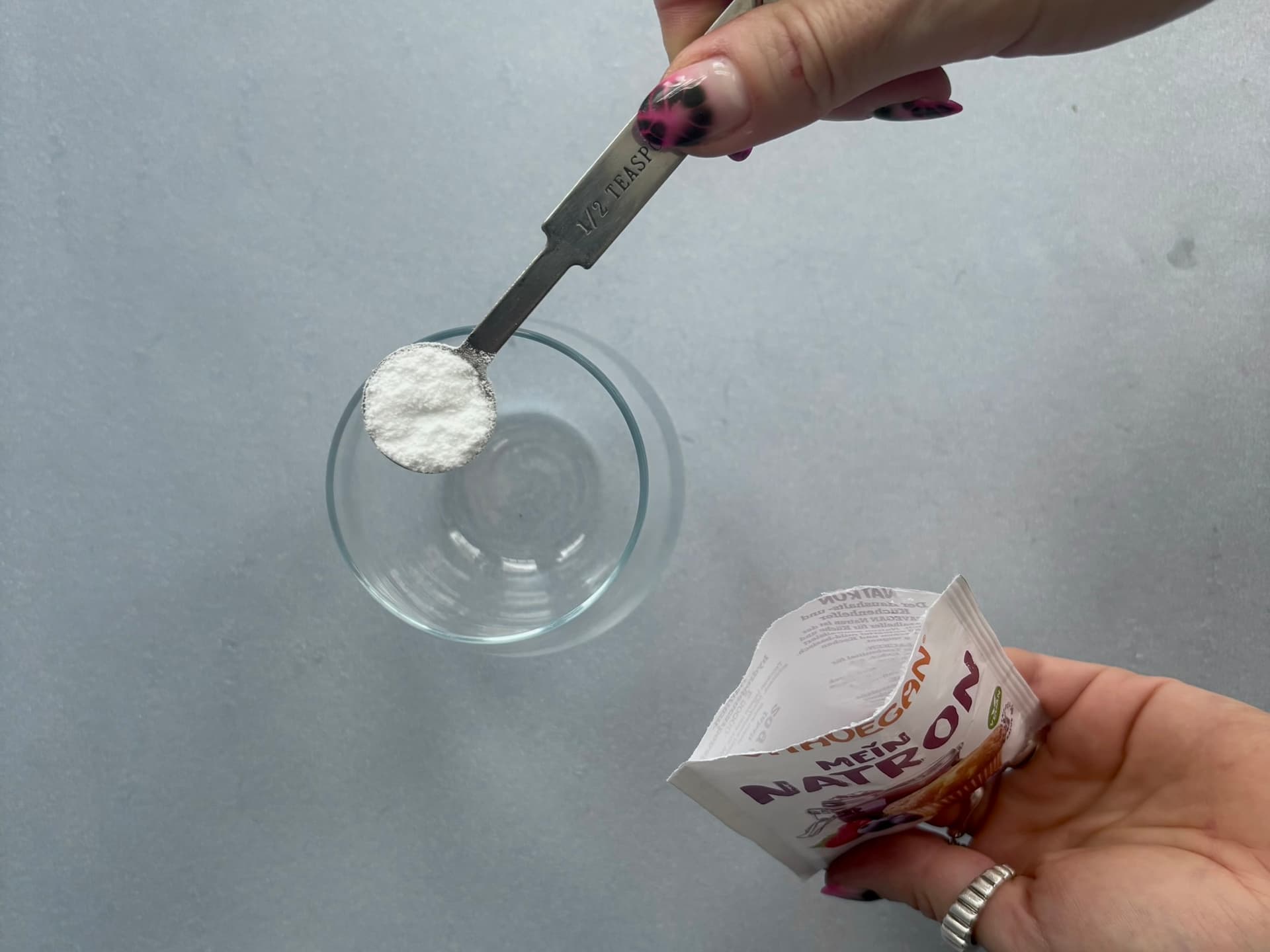 A hand pours half a teaspoon of baking powder from a packet into a clear glass bowl on a gray surface.