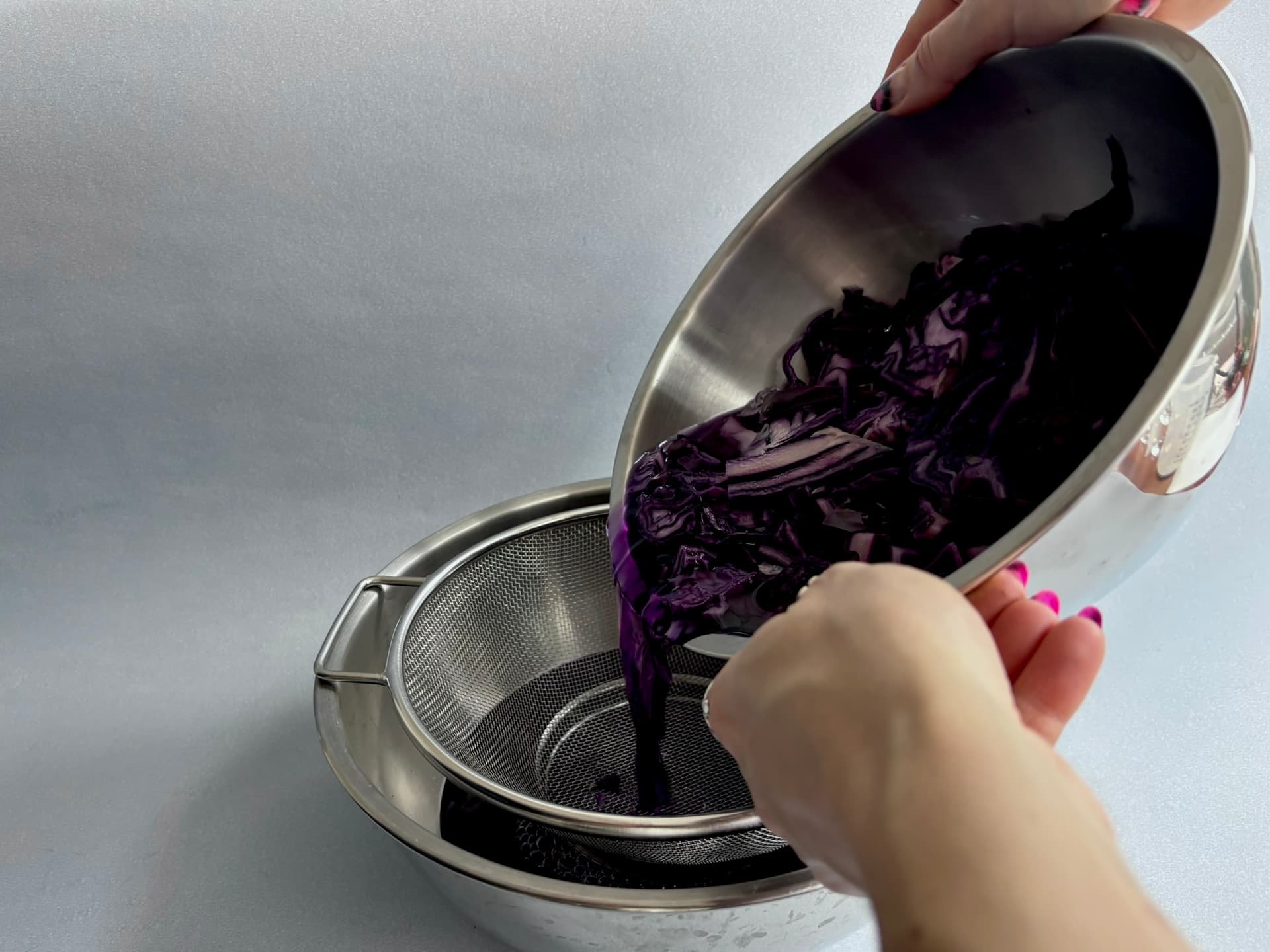 Hands pouring shredded purple cabbage from a bowl into a strainer over another bowl.