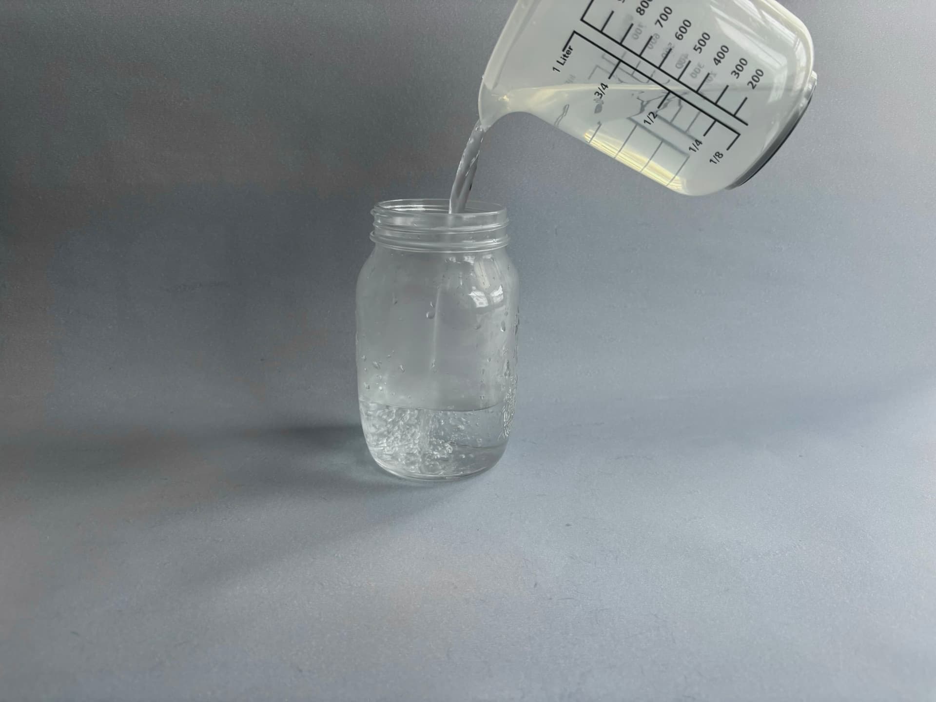 A measuring jar pours water into a glass mason jar on a gray surface.