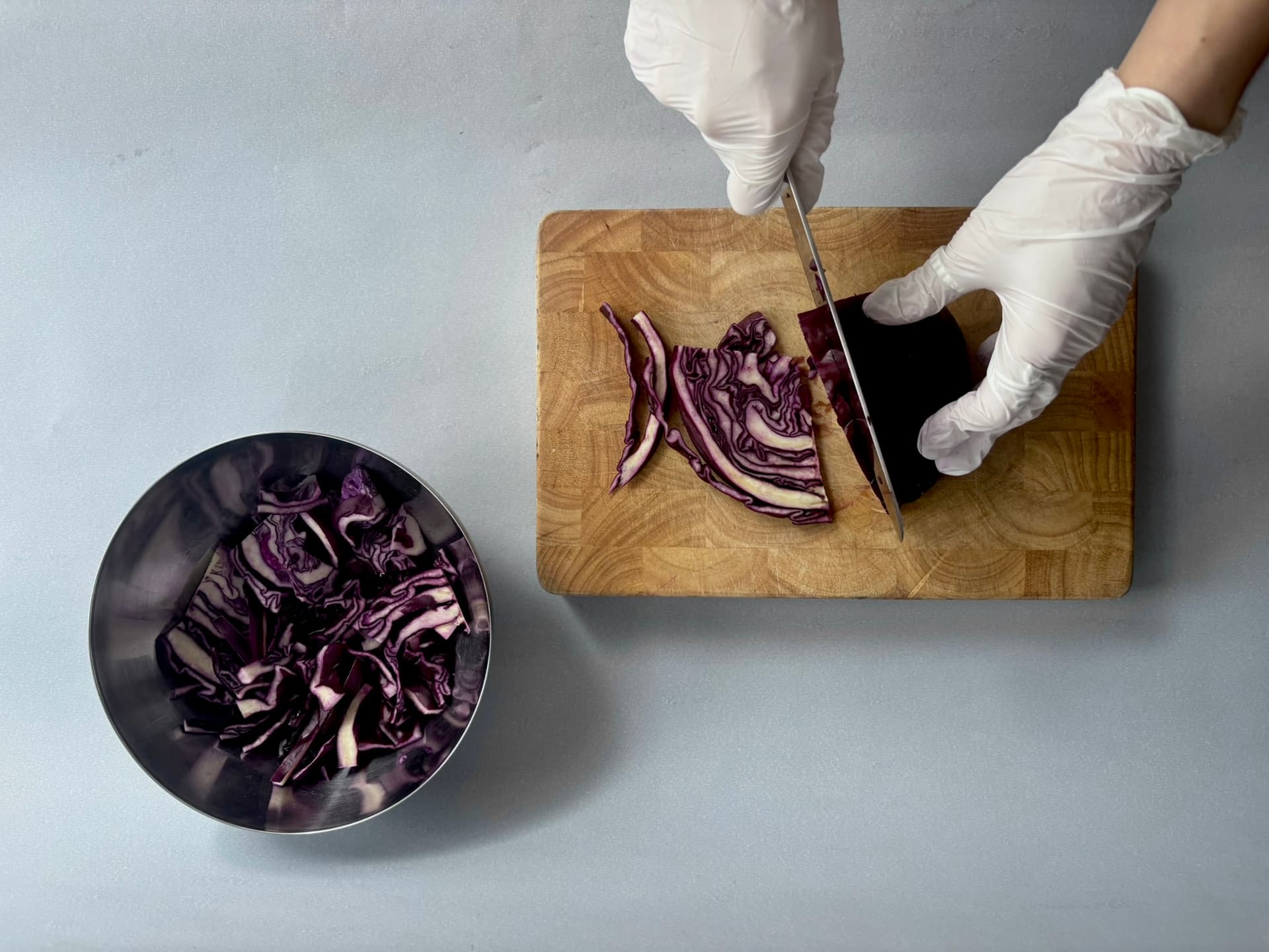 Gloved hands slicing purple cabbage on a wooden board, with a metal bowl containing chopped cabbage beside it.
