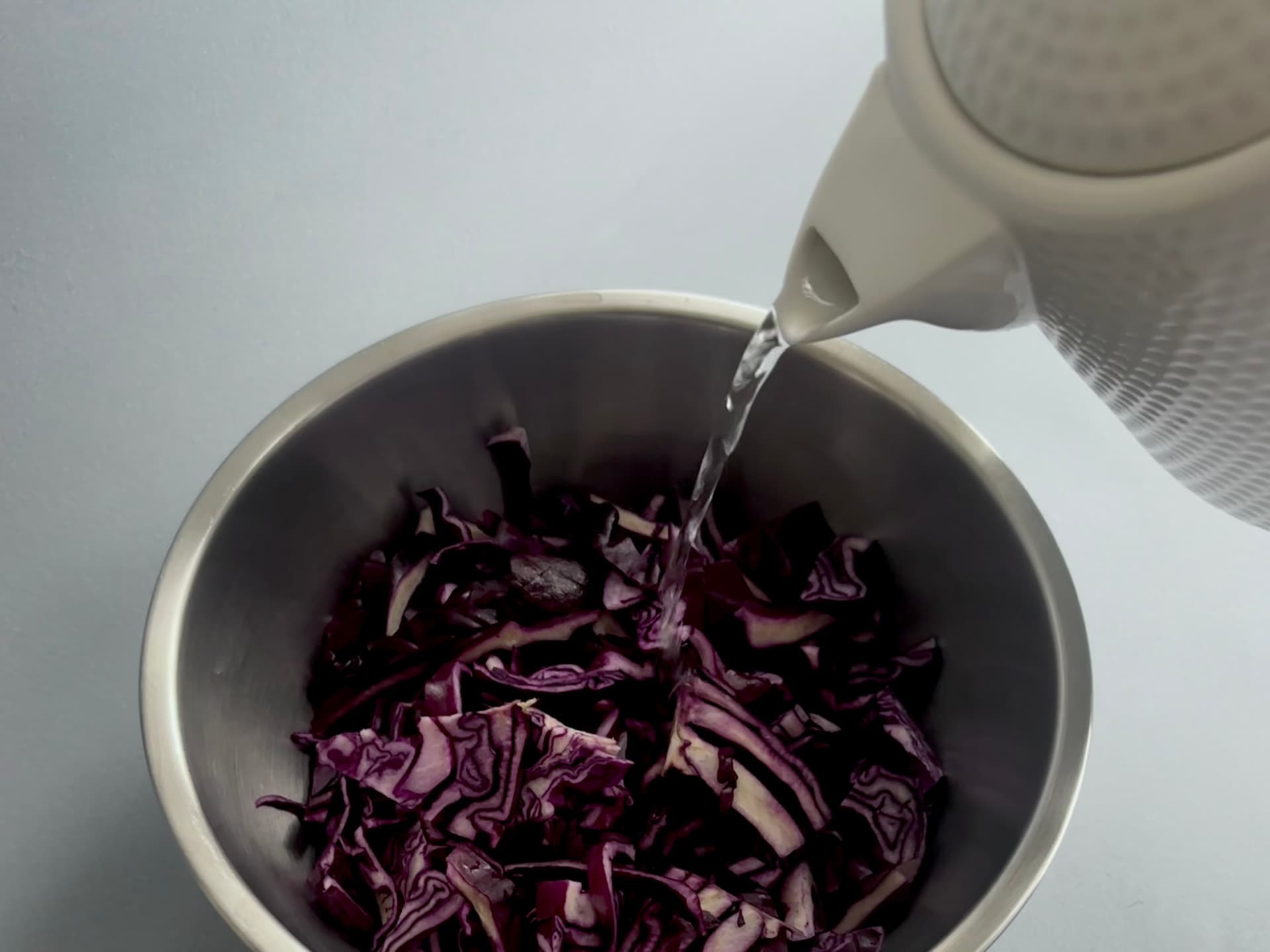 Hot water being poured from a kettle into a metal bowl containing chopped purple cabbage.