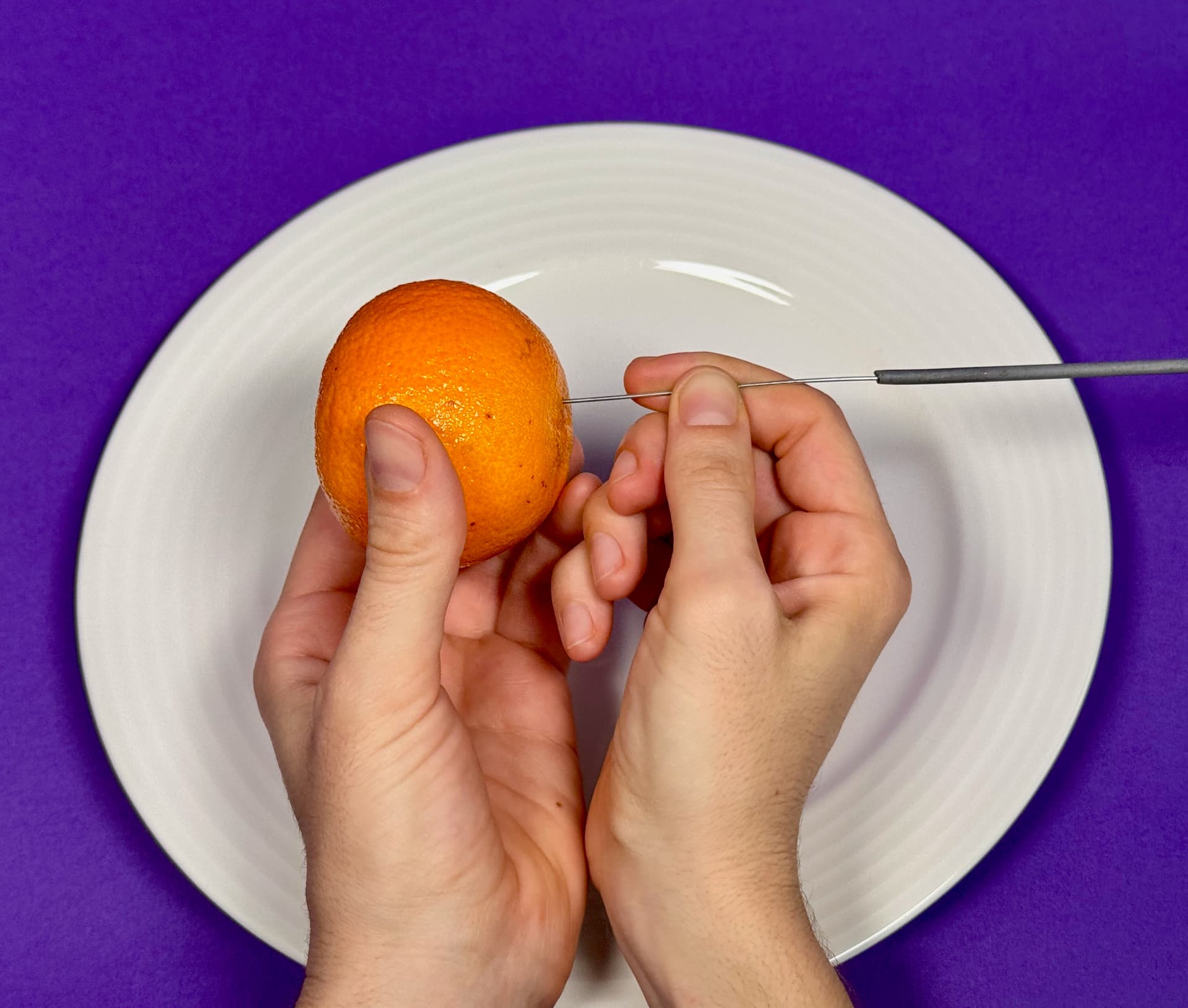Hände, die eine Orange mit einem Messer über einem weißen Teller auf violettem Hintergrund schälen.
