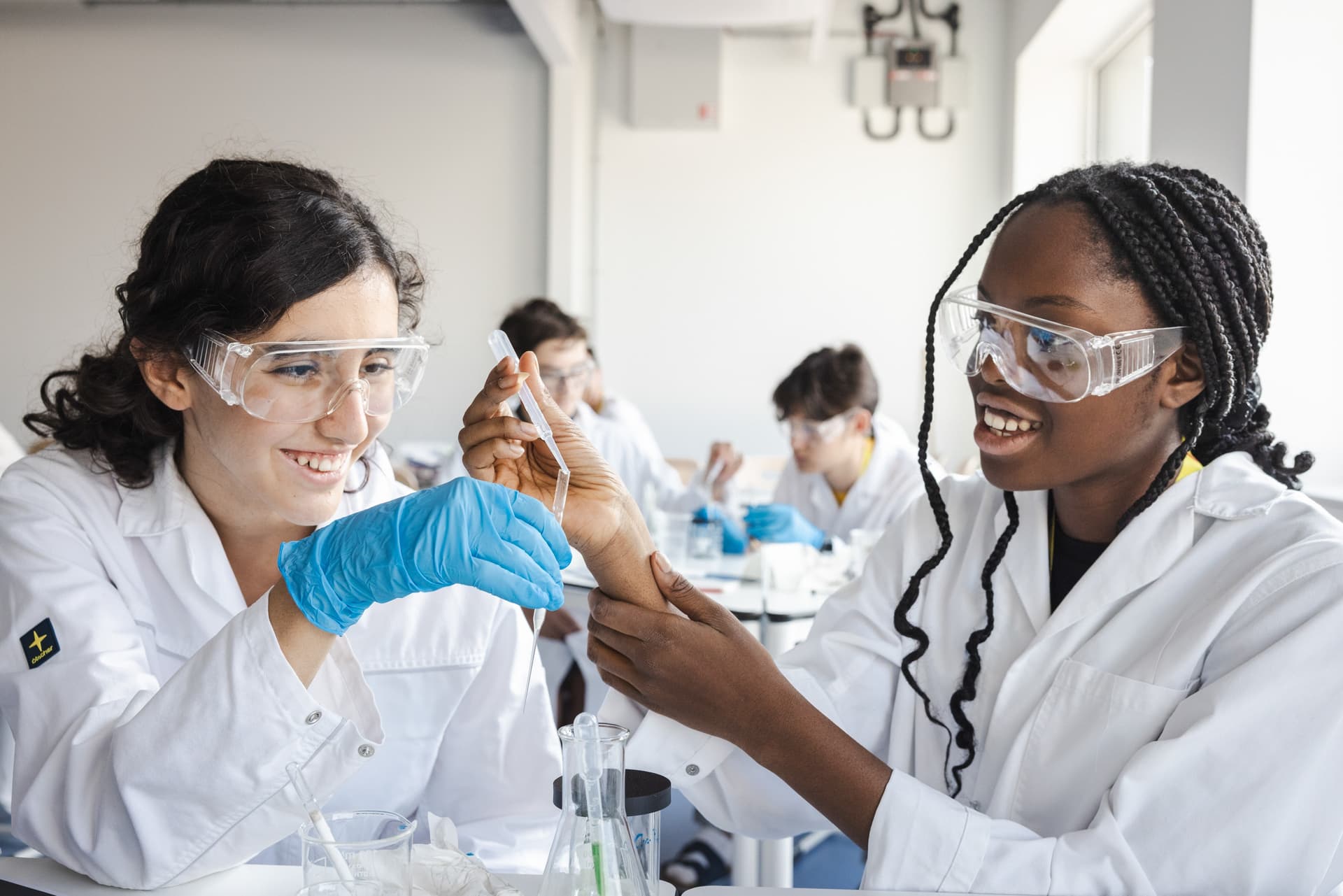 Zwei junge Frauen in Laborkitteln und Schutzbrillen arbeiten gemeinsam an einem wissenschaftlichen Experiment. Eine hält eine Pipette, während die andere ihre Hand unterstützt. Beide lachen und wirken motiviert. Im Hintergrund sitzen weitere junge Menschen in ähnlicher Schutzkleidung an Labortischen.