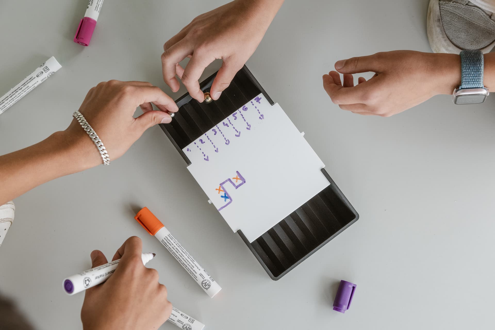 Hände, die mit bunten Stiften auf Papier zeichnen, umgeben von einem Tablett und verstreuten Stiften auf einer weißen Fläche.