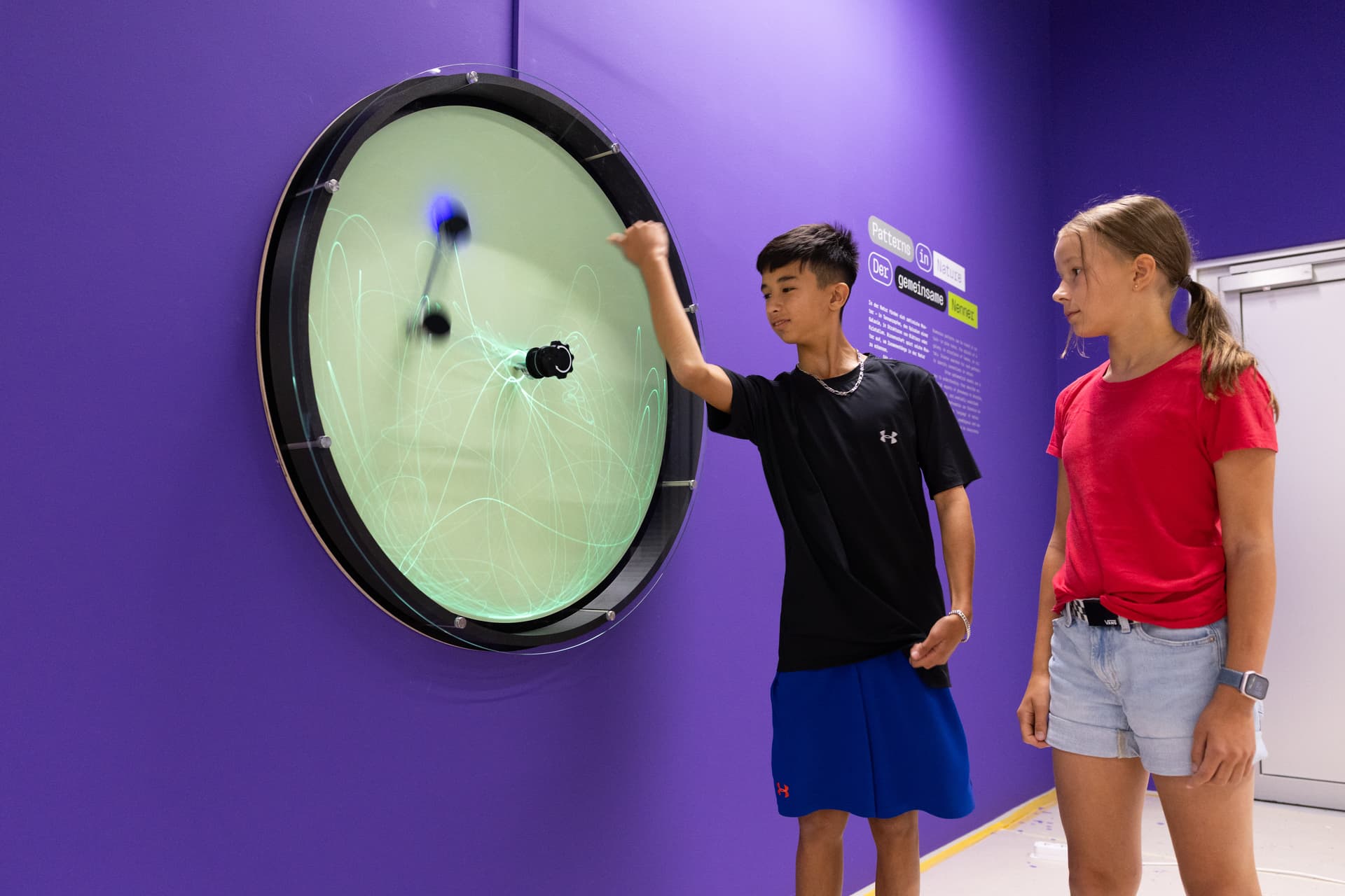 Zwei Personen interagieren mit einer großen kreisförmigen Lichtanzeige an einer violetten Wand und zeichnen mit einem Stift Muster.