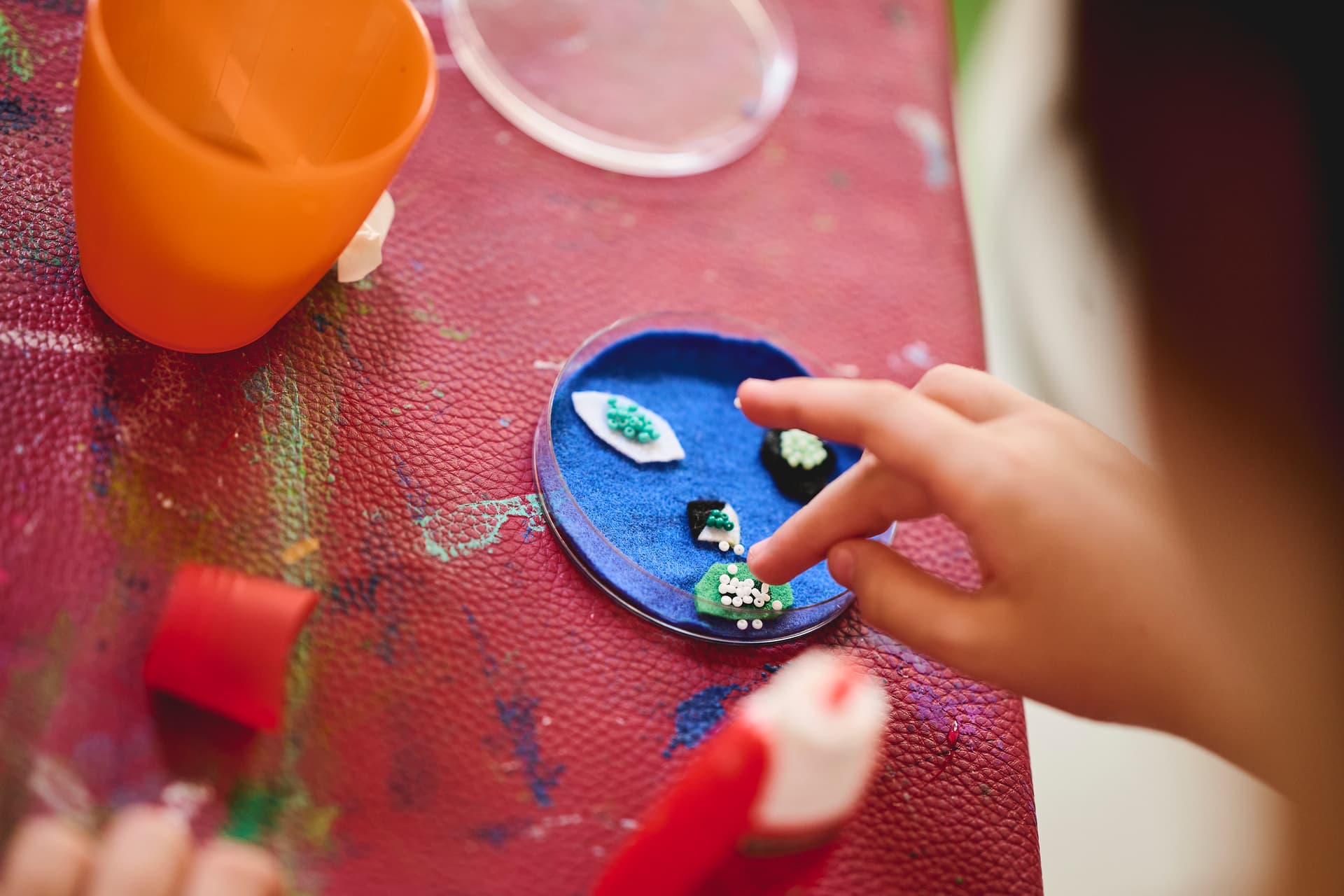 Kinderhand, die kleine dekorative Elemente auf einem blauen Tablett auf einer strukturierten roten Oberfläche mit bunten Farbspritzern anordnet; daneben steht ein orangefarbener Becher.