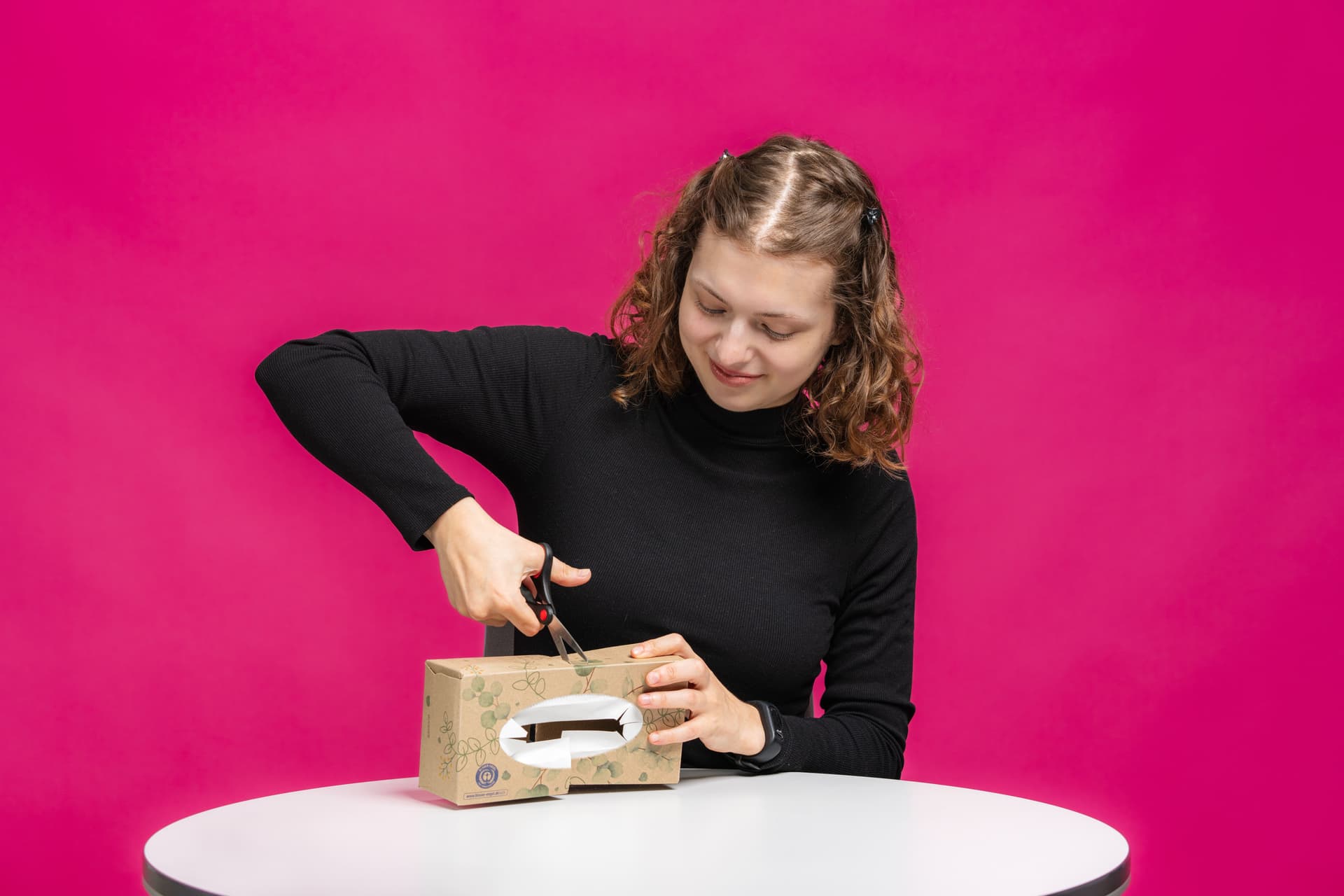 Eine Person in einem schwarzen Pullover schneidet mit einer Schere einen Karton auf einem weißen Tisch vor einem leuchtend pinkfarbenen Hintergrund auf.