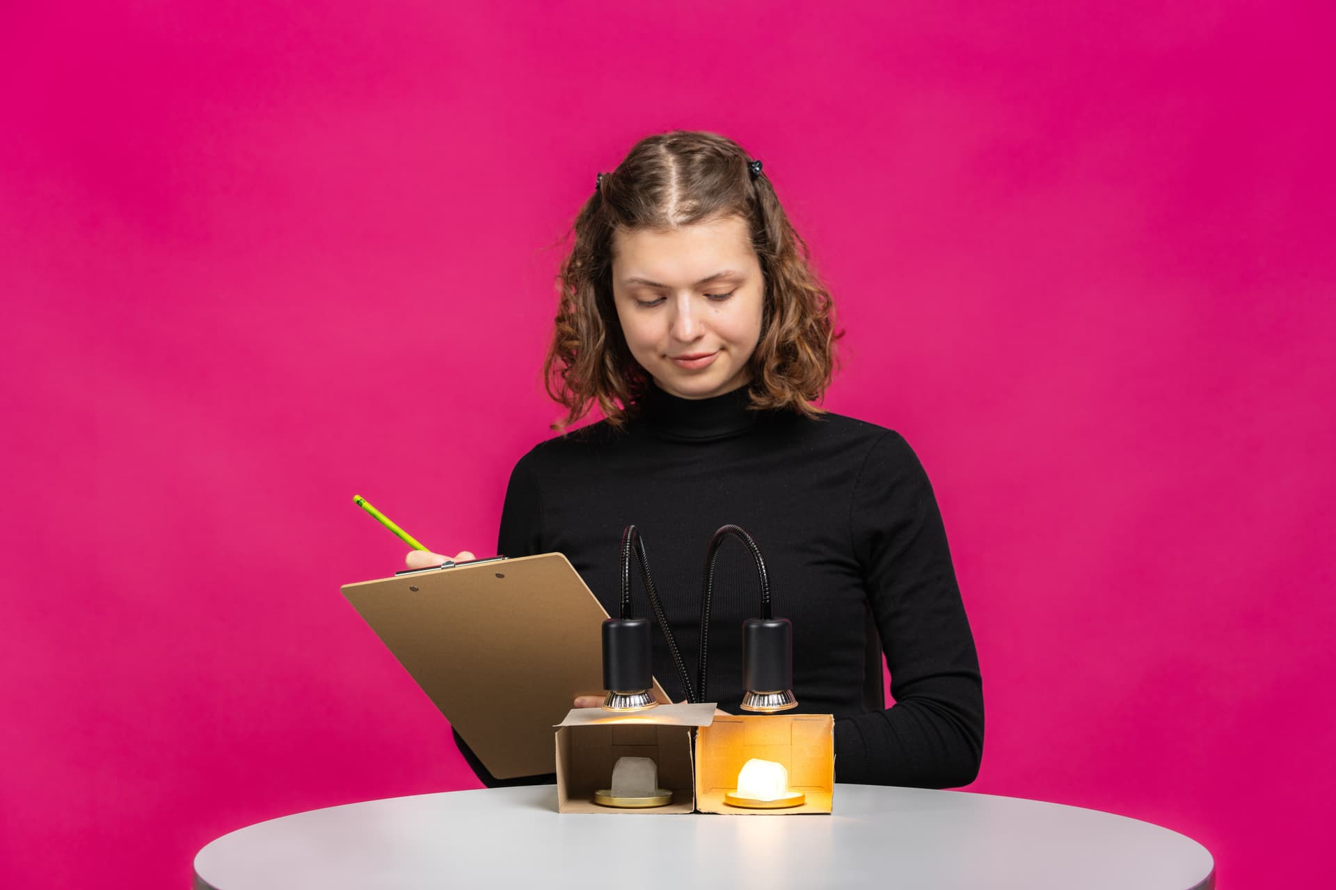 Eine Frau in einem schwarzen Oberteil, die zwei brennende Lampen auf einem Tisch begutachtet, hält ein Klemmbrett und einen Bleistift in der Hand, vor einem rosa Hintergrund.