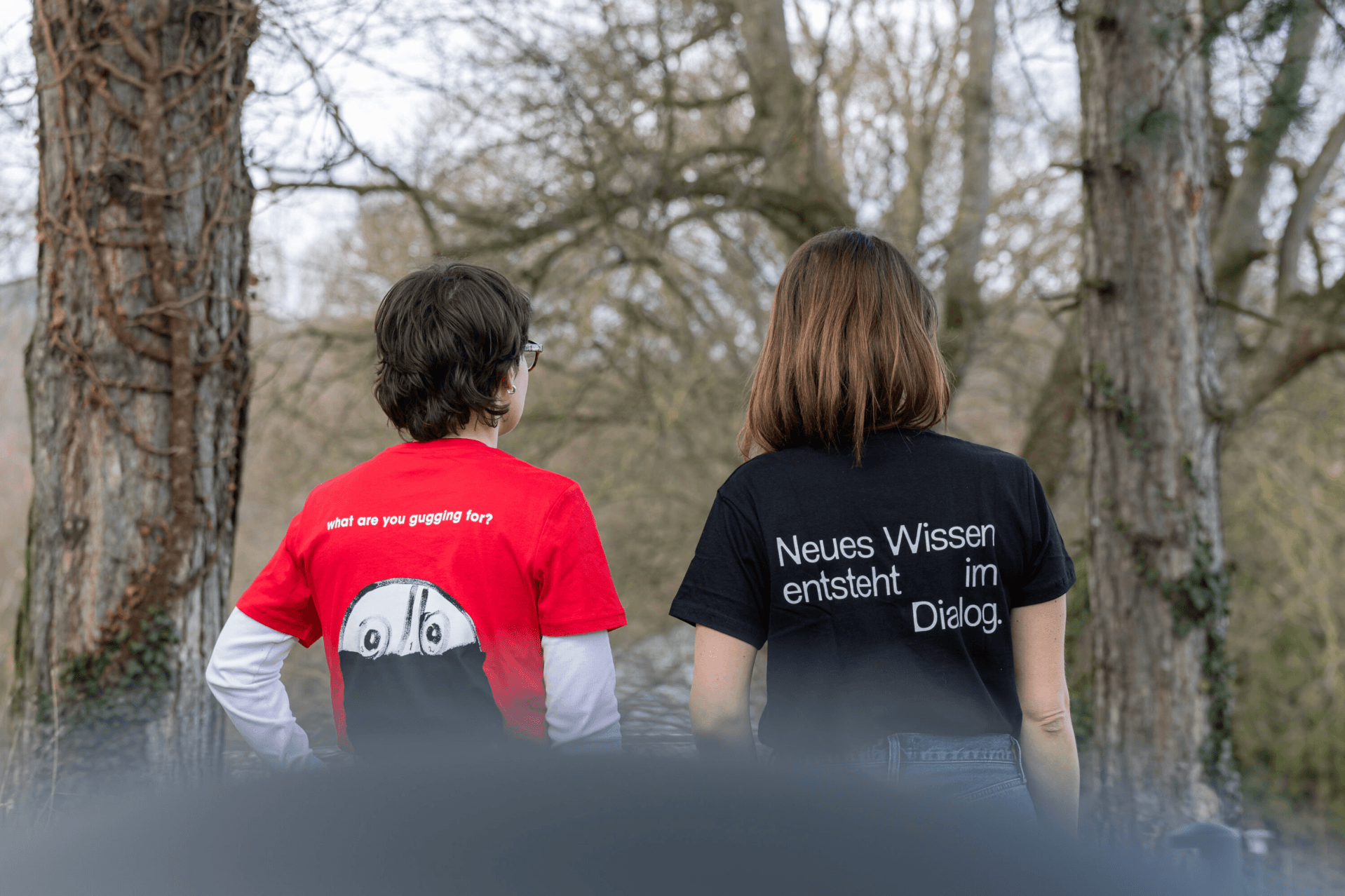 Zwei Personen stehen im Freien, von hinten gesehen, und tragen T-Shirts mit Aufdruck. Das linke T-Shirt ist rot, das rechte schwarz. Im Hintergrund sind Bäume zu sehen.