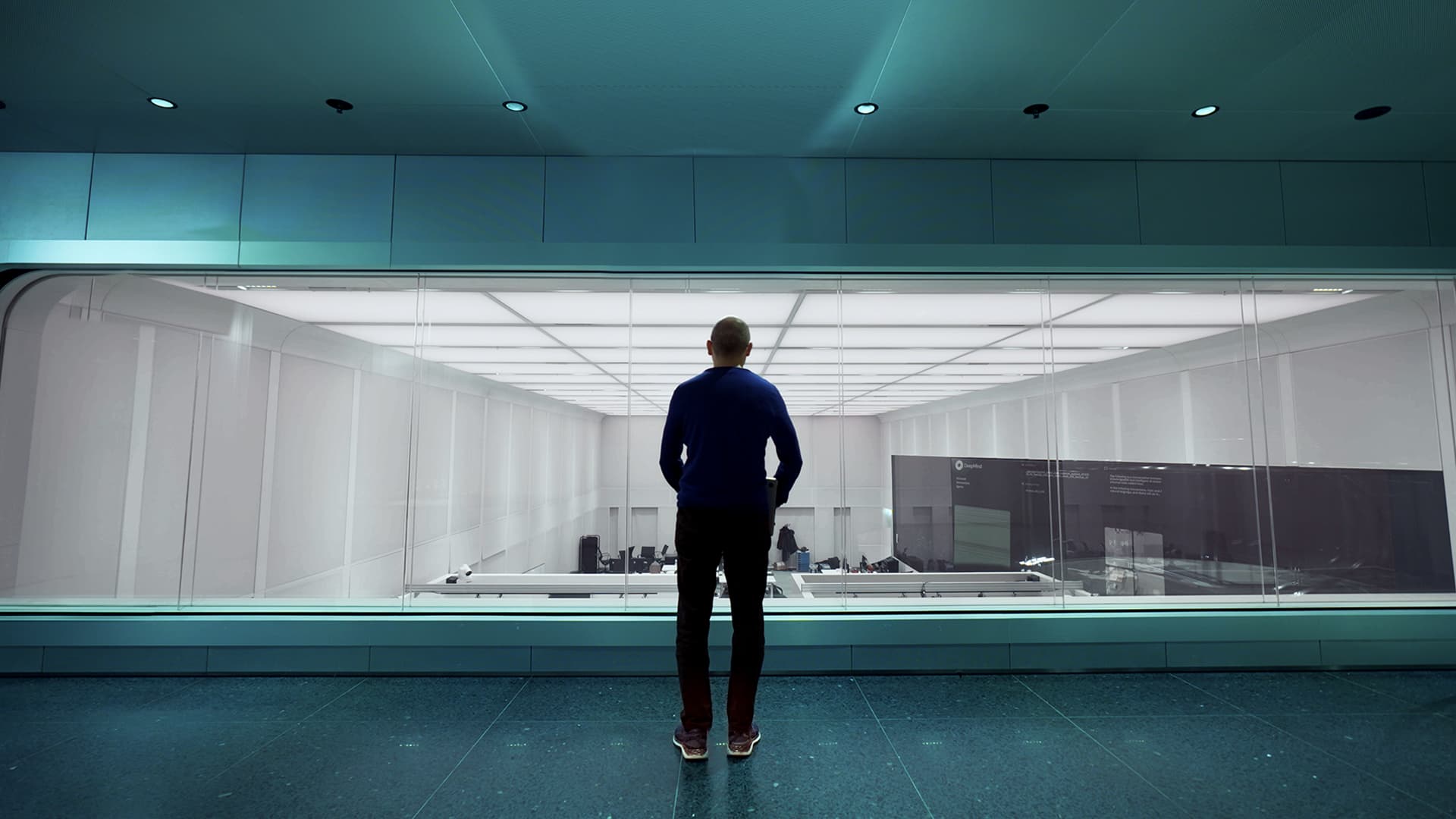 A person stands in a modern room, viewing a large glass window revealing an empty, futuristic space.