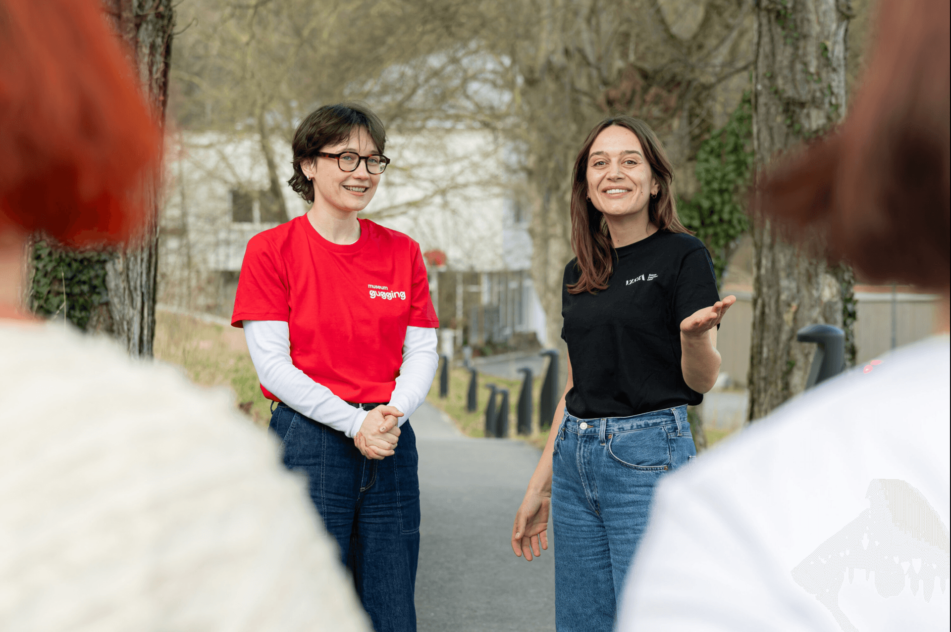 Zwei Frauen in Freizeitkleidung, die lächelnd miteinander sprechen, während sie auf einem von Bäumen gesäumten Weg stehen; im Vordergrund sind verschwommene Menschen zu sehen.