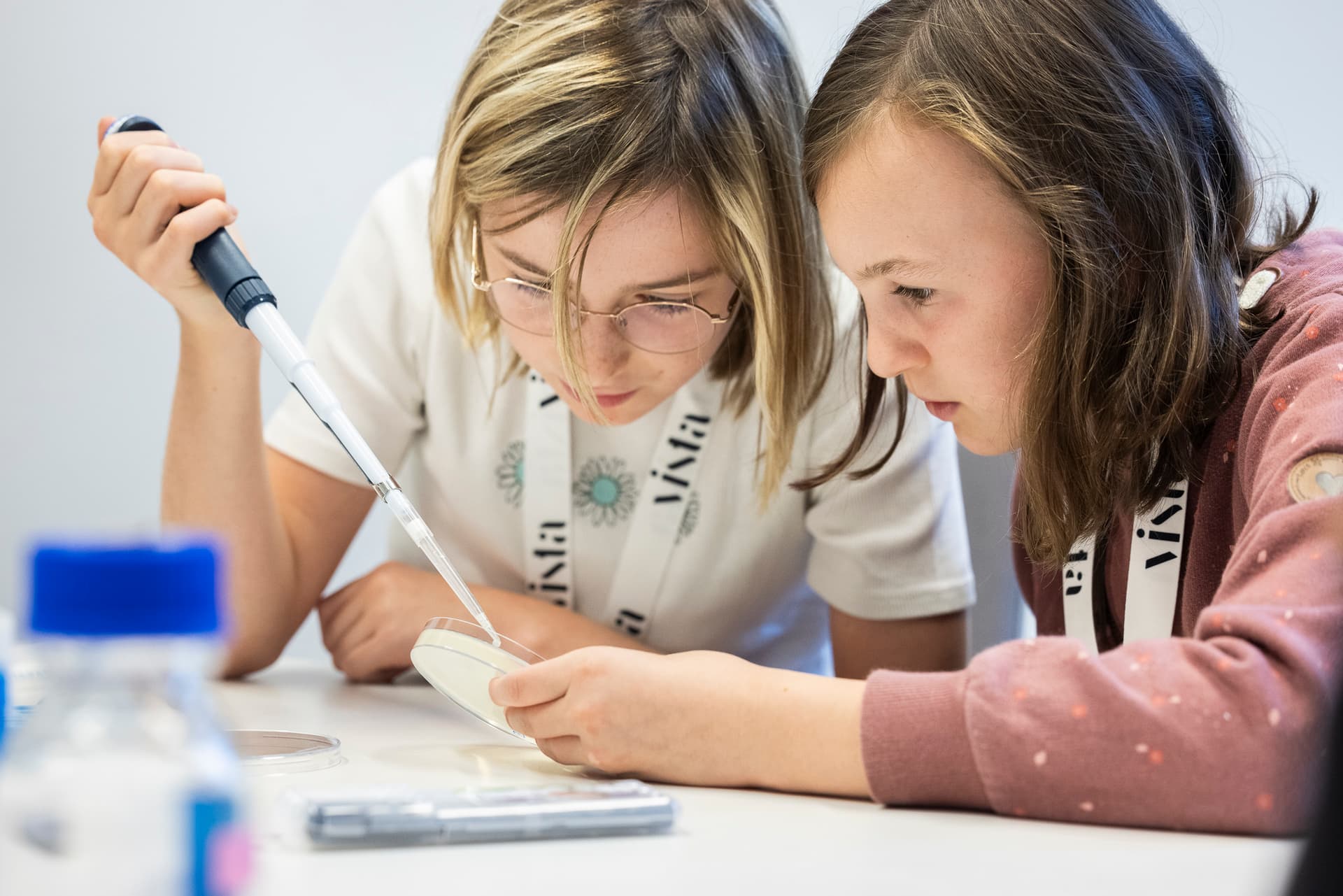 Zwei Mädchen untersuchen in einem Labor mit einer Pipette aufmerksam eine Petrischale. Sie wirken konzentriert und vertieft in ein wissenschaftliches Experiment.