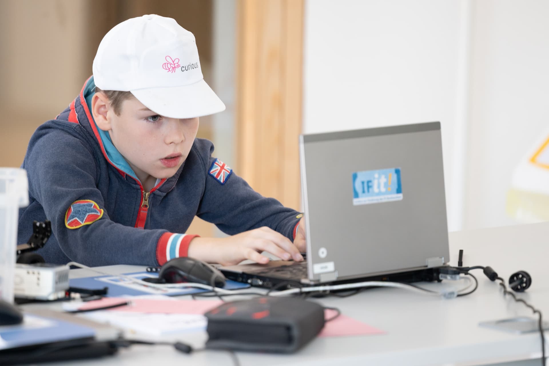 Ein kleiner Junge mit einer weißen Mütze arbeitet konzentriert an einem Laptop an einem Tisch, umgeben von verschiedenen Dokumenten und Geräten.