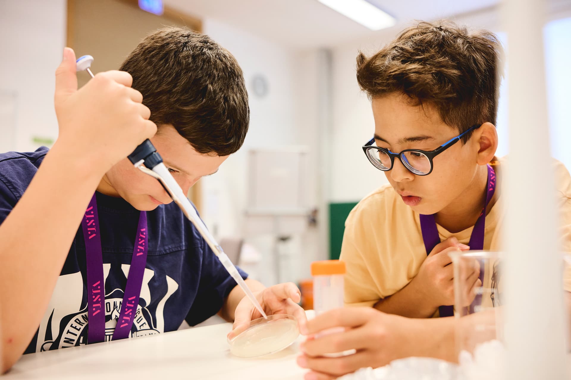 Two boys conducting a science experiment, using pipettes and petri dishes in a classroom setting.