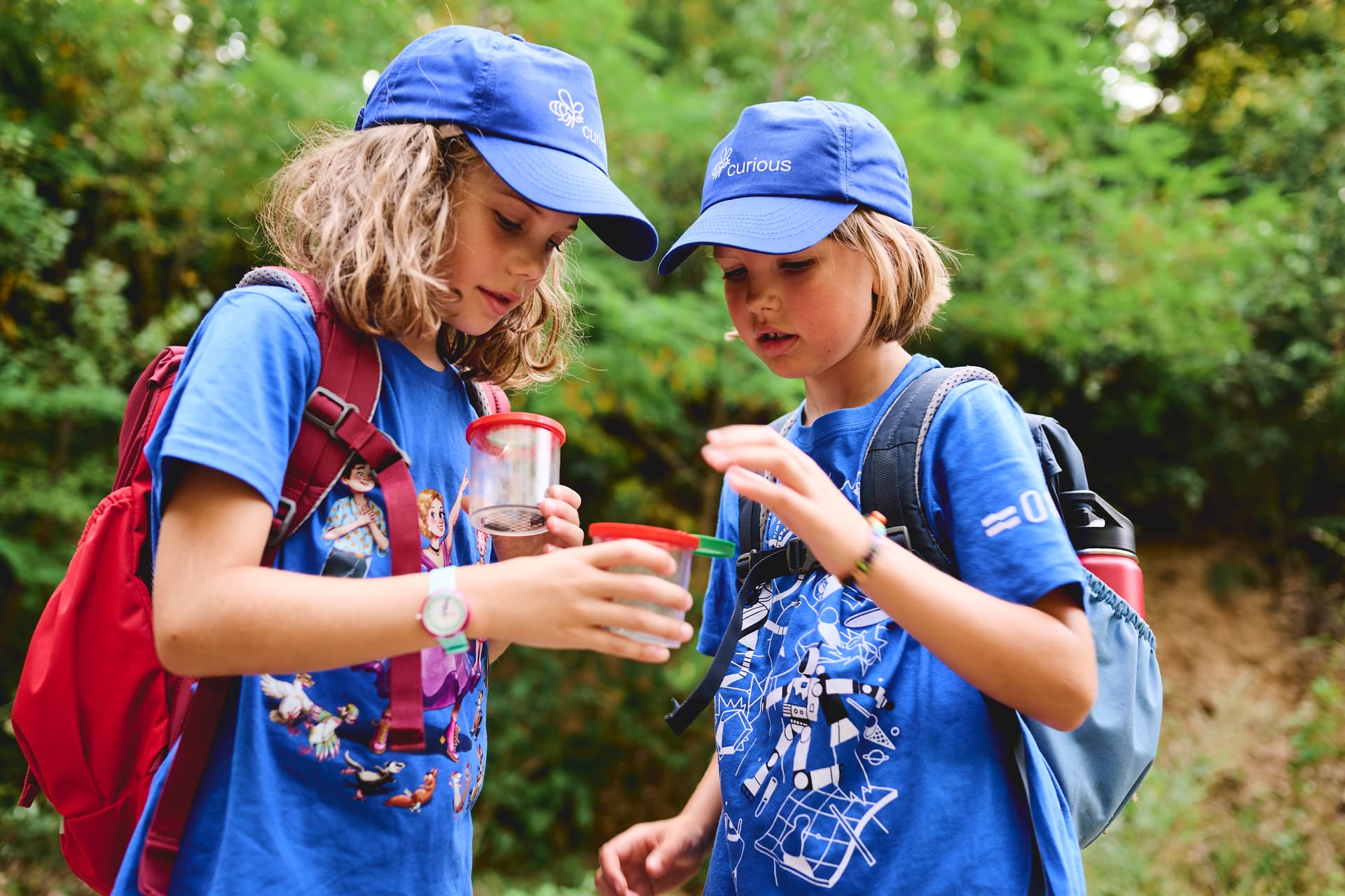 Two children wearing blue caps and backpacks examine a plastic jar in a forest setting.