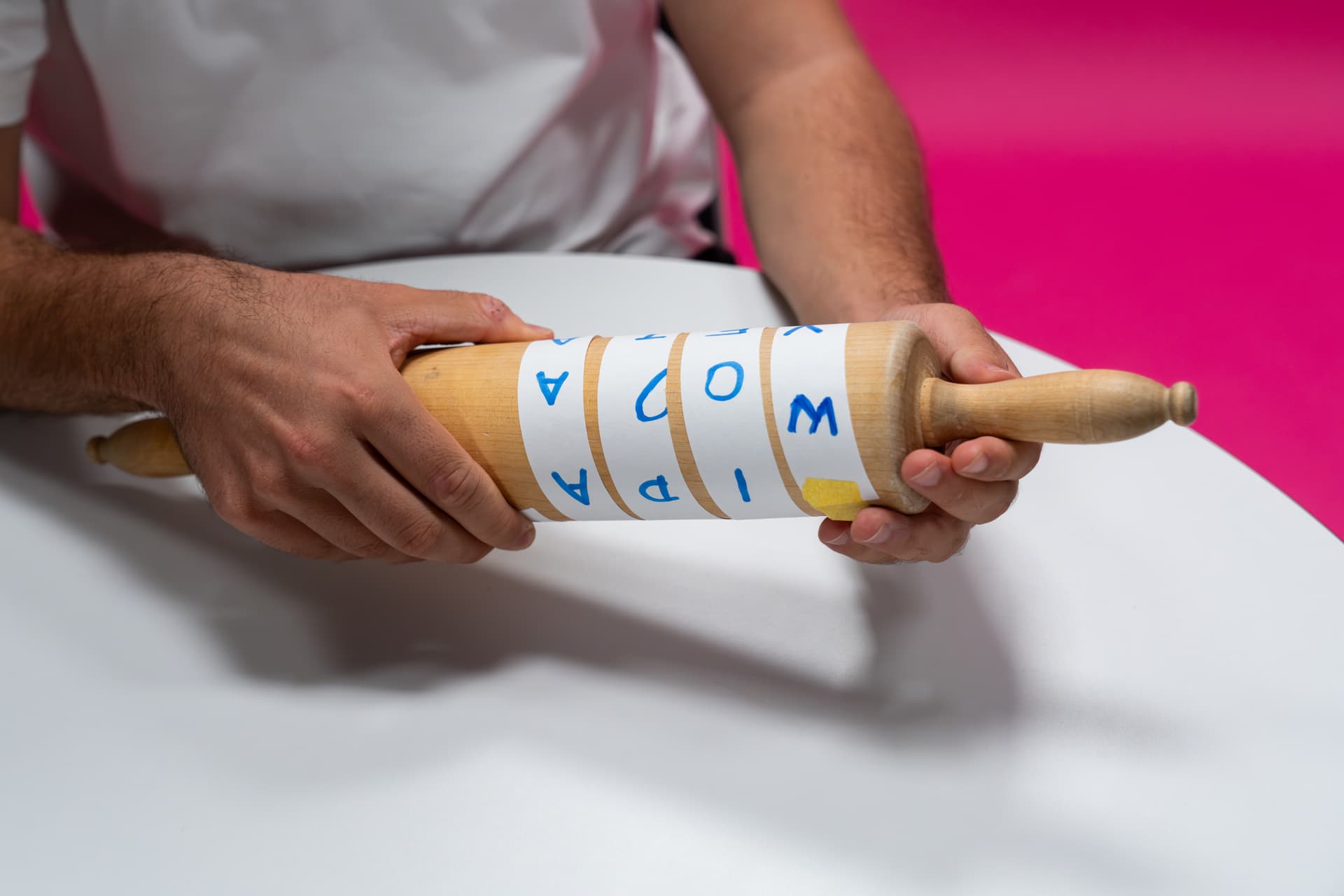 Eine Person, die ein mit Papier mit blauen Buchstaben umwickeltes Holzrollbrett in der Hand hält und an einem runden weißen Tisch vor einem rosa Hintergrund sitzt.