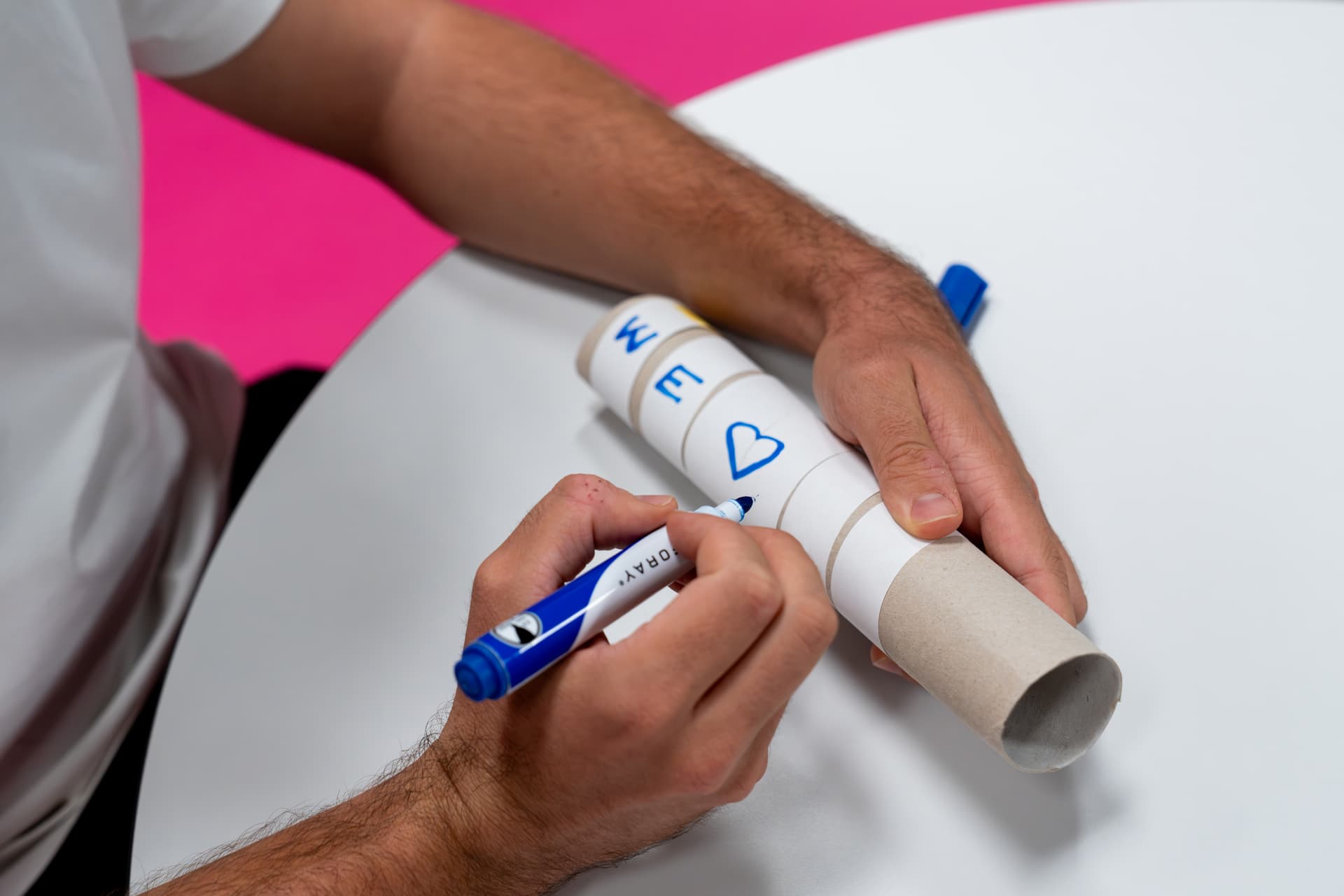 Eine Person, die mit einem blauen Filzstift auf einem zylindrischen Karton schreibt, der mit Buchstaben und Symbolen verziert ist, auf einem runden weißen Tisch.