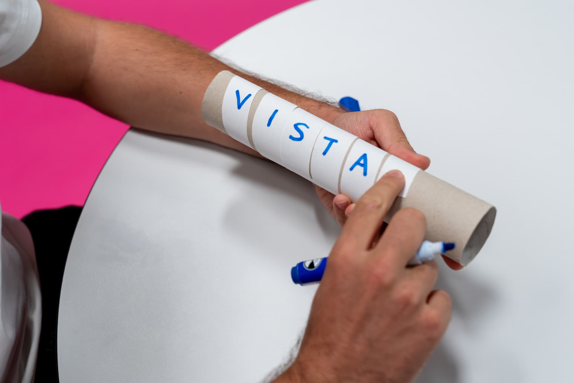 Eine Person, die eine Papprolle in der Hand hält, um die ein Zettel mit der Aufschrift „VISTA“ gewickelt ist, sitzt an einem weißen runden Tisch vor einem rosa Hintergrund.
