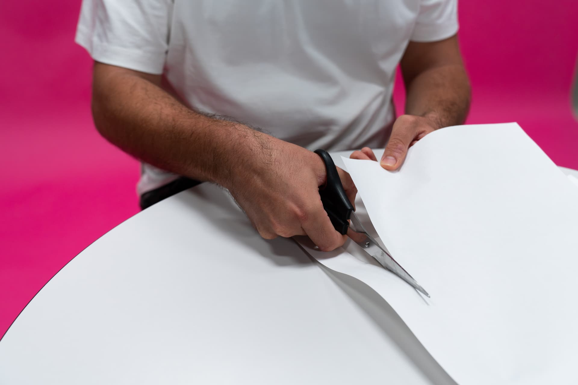 Person, die vor einem pinken Hintergrund weißes Papier mit einer Schere schneidet.