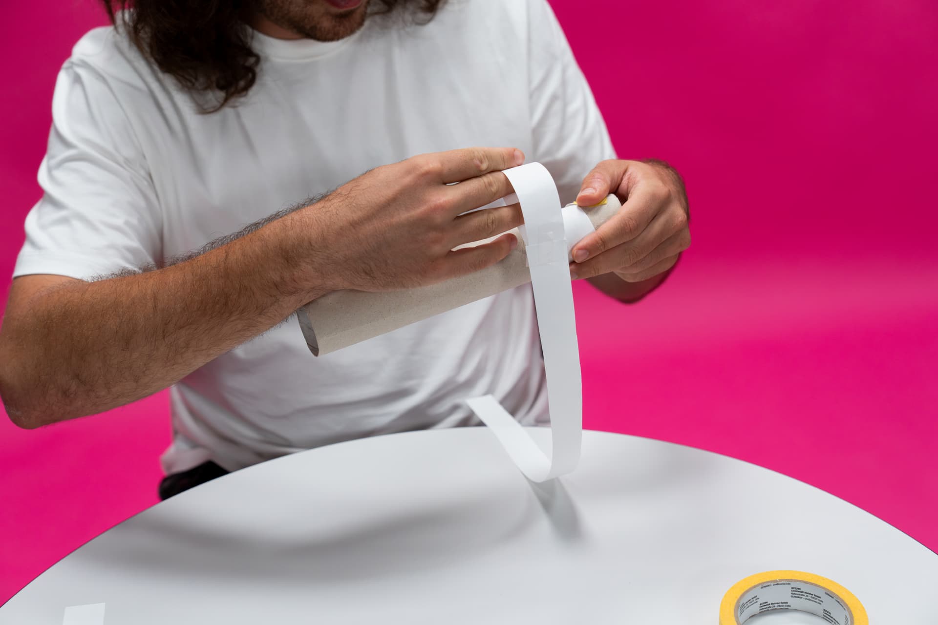 Eine Person in einem weißen Hemd wickelt Klebeband um eine Papprolle. Der Tisch ist weiß, der Hintergrund ist rosa, und auf dem Tisch liegt eine Rolle Klebeband.